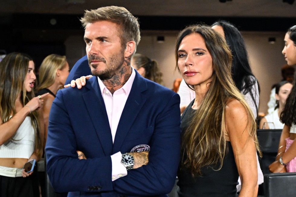 David Beckham and Victoria Beckham David Beckham and Victoria Beckham. David has his arms folded looking off to the side and Victoria has her arm around his shoulder looking to camera.